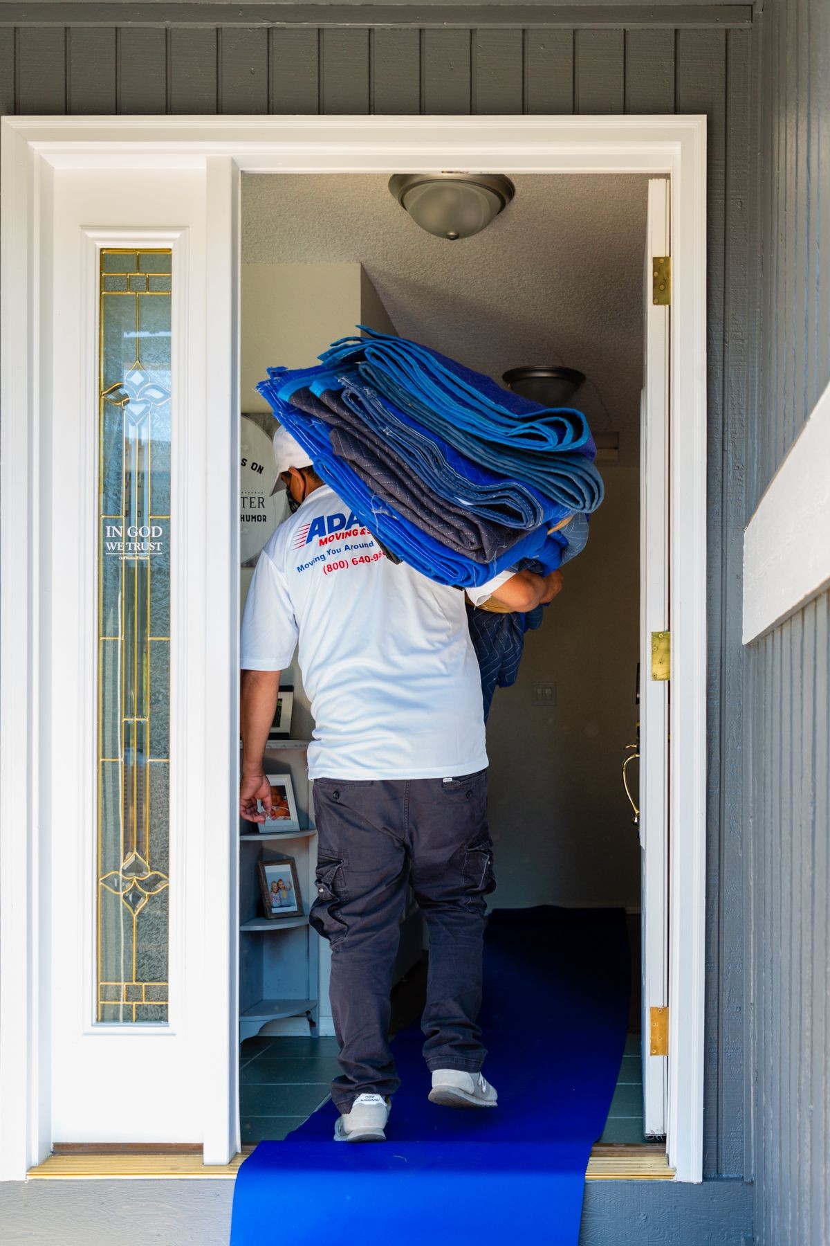 Person carrying folded moving blankets into a house, standing on a blue protective floor covering.