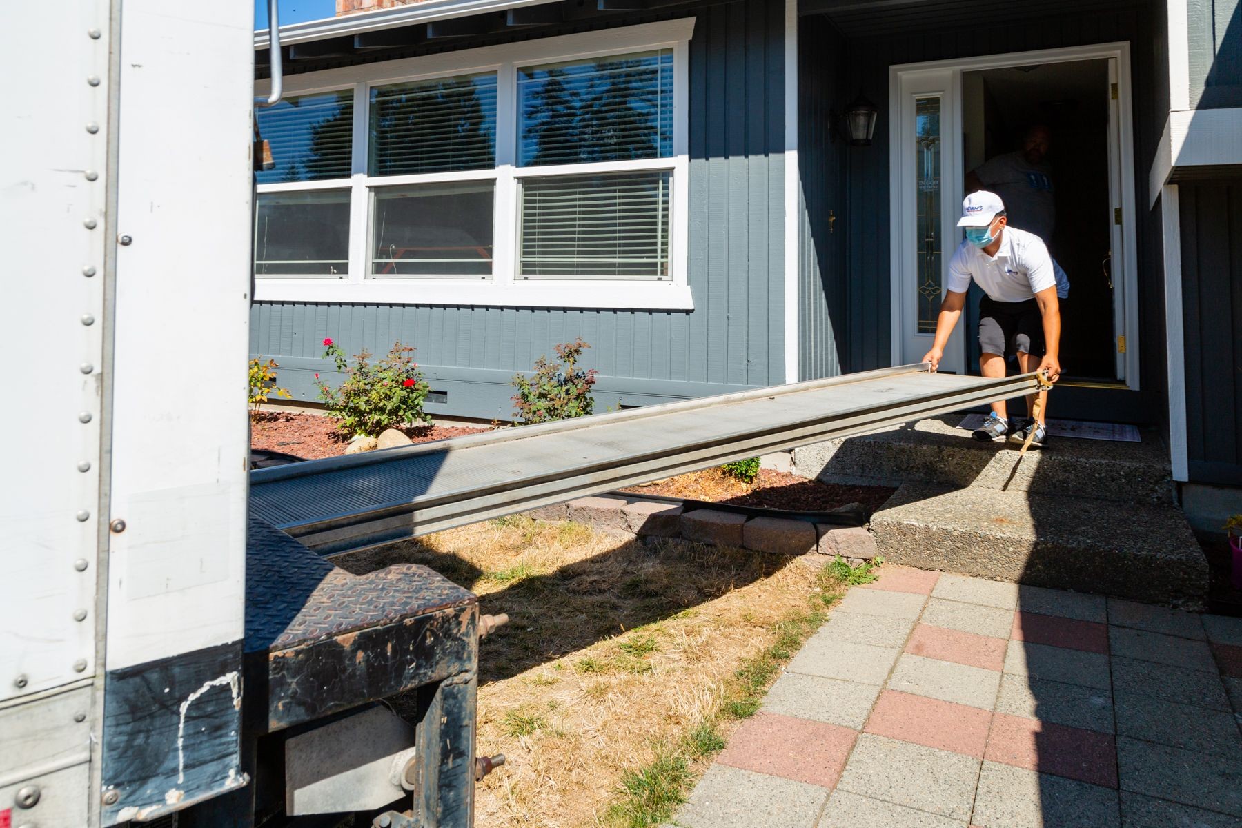 Person in white clothes setting up a ramp from a truck to the entrance of a blue house.